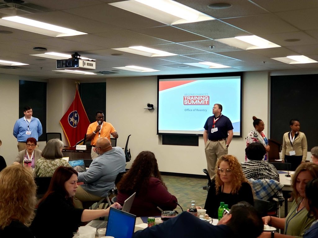 A trainer stands in front of the Tennessee state flag and presents to a large room of people seated at round tables