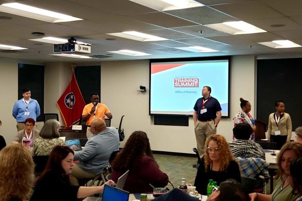 A trainer stands in front of the Tennessee state flag and presents to a large room of people seated at round tables